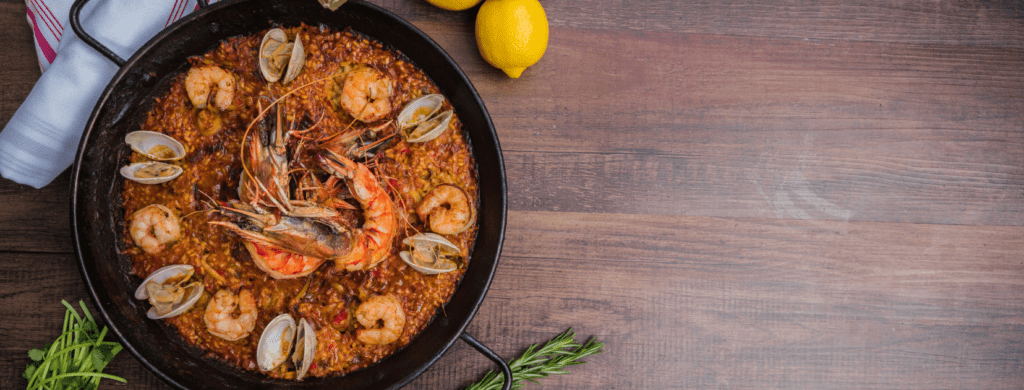 Overhead view of seafood paella in a black paella pan with shrimp, clams, lemon, rosemary, and herbs on a wooden table.