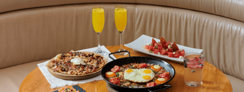 Bulla Gastrobar brunch spread featuring flatbread, a skillet dish with eggs, watermelon salad, mimosas, and a refreshing cocktail on a round table.