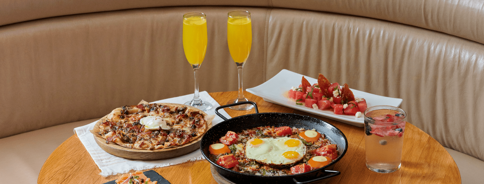 Bulla Gastrobar brunch spread featuring flatbread, a skillet dish with eggs, watermelon salad, mimosas, and a refreshing cocktail on a round table.
