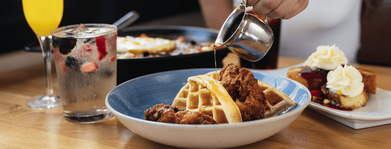 Bulla Gastrobar brunch spread featuring chicken and waffles with syrup, a refreshing cocktail, mimosa, skillet eggs, and dessert on a wooden table.