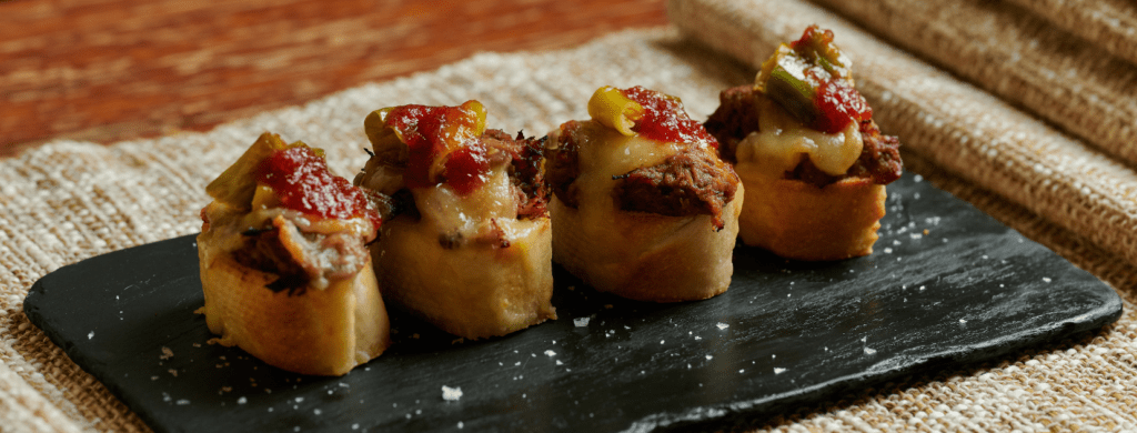 Happy hour tapas menu served on a black slate plate, featuring toasted bread topped with savory meat, melted cheese, peppers, and red sauce.
