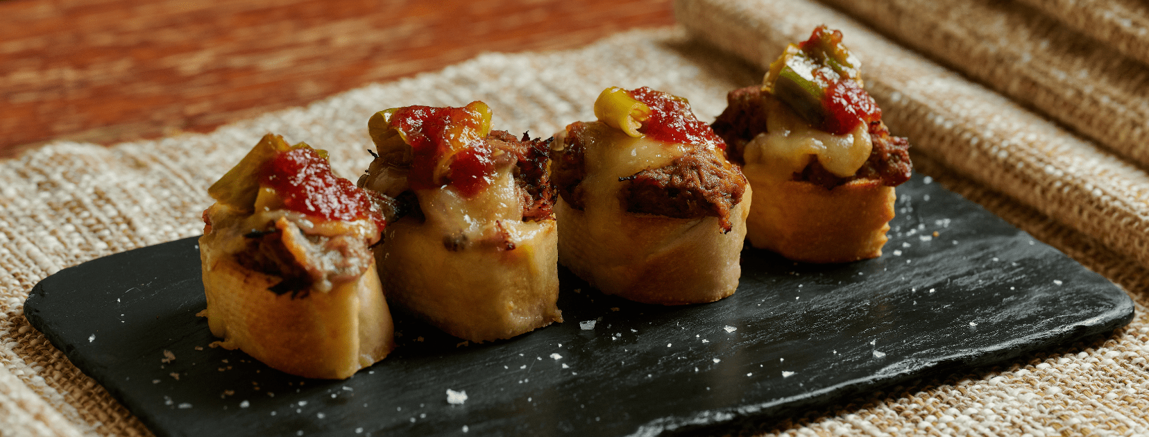 Happy hour tapas menu served on a black slate plate, featuring toasted bread topped with savory meat, melted cheese, peppers, and red sauce.