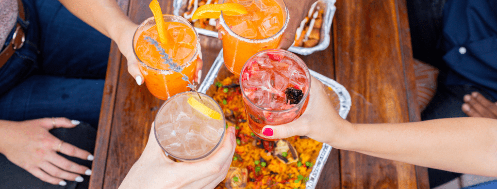 Bulla Gastrobar catering spread featuring guests toasting with colorful cocktails over a large paella tray on a wooden table.
