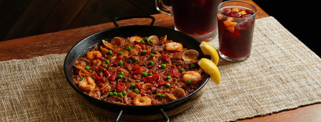 Seafood paella served in a black paella pan with shrimp, clams, peas, roasted peppers, lemon wedges, and glasses of red sangría on a wooden table.